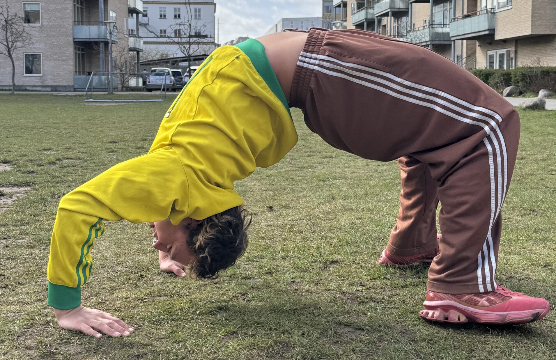 Foto de Agustín haciendo un ejercicio de gimnasia (puente) en un pasto al aire libre con edificios atrás y un cielo azul.